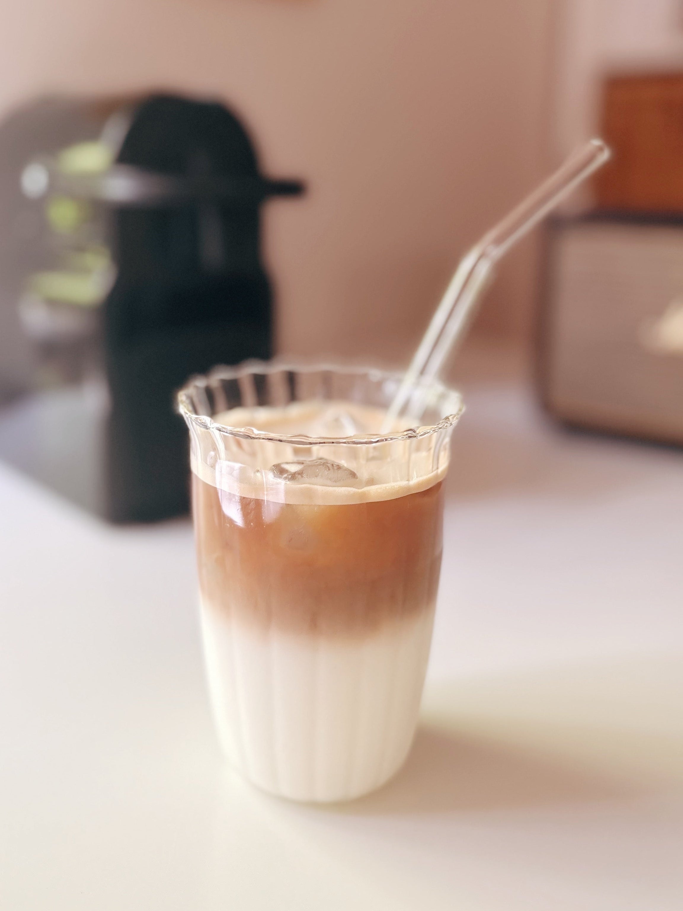 A clear ribbed glass cup filled with an iced coffee drink layered in dark brown and creamy white, with a clear bent straw, set on a white tabletop with a blurred coffee maker and speaker in the background.