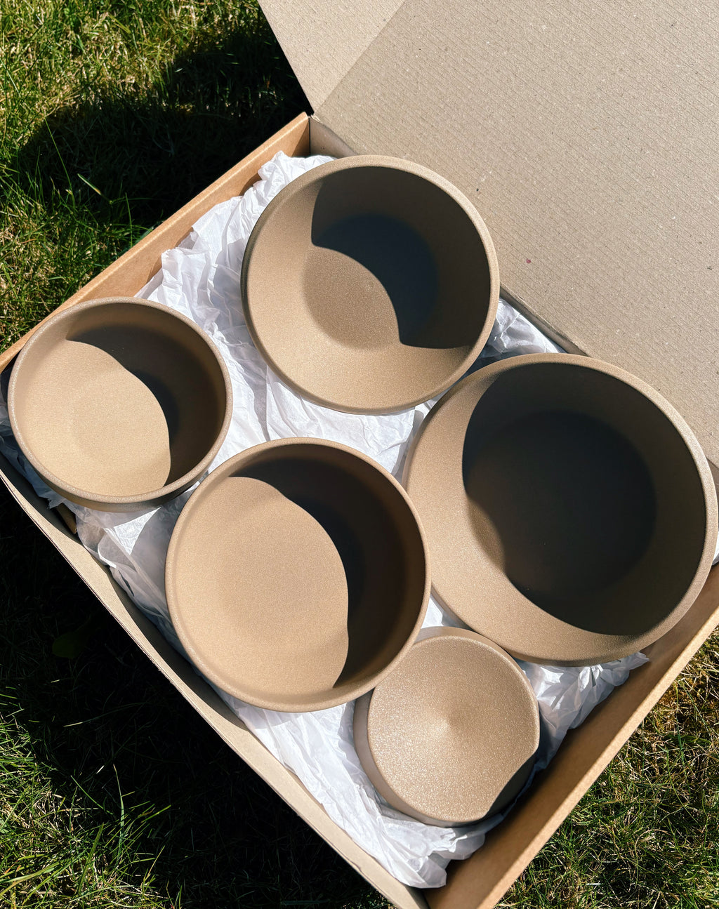 Five matte beige ceramic bowls of varying sizes arranged inside a cardboard box with white paper packing material, photographed outdoors on grass in bright natural sunlight.