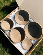 Five matte beige ceramic bowls of varying sizes arranged inside a cardboard box with white paper packing material, photographed outdoors on grass in bright natural sunlight.