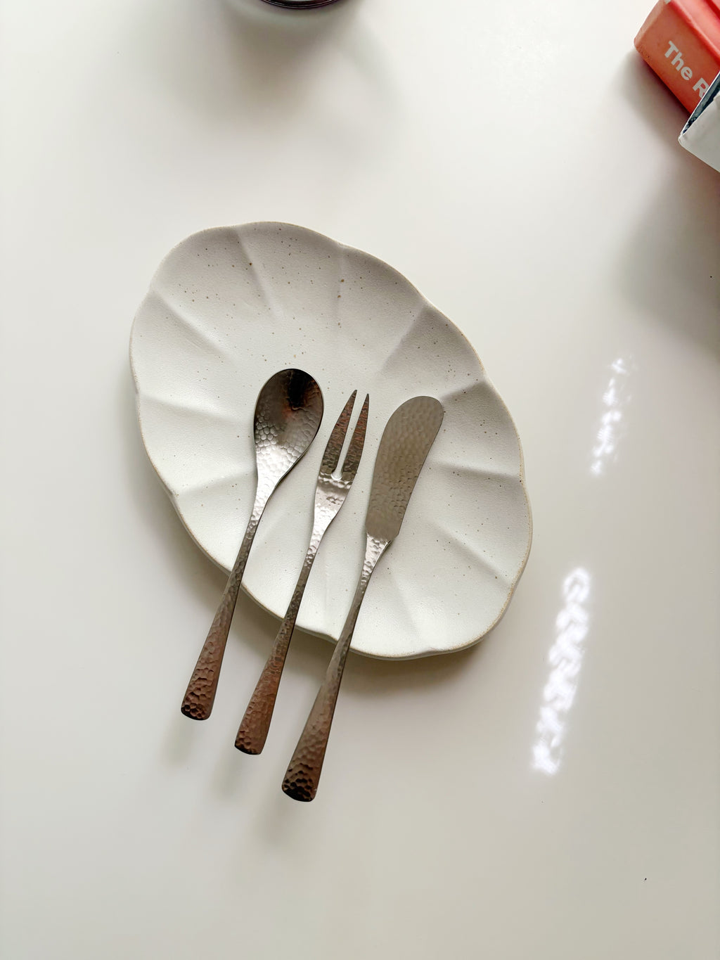 Three hammered metal utensils—a spoon, a fork, and a knife—rest on a scalloped off-white ceramic plate against a clean white background.