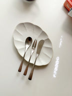 Three hammered metal utensils—a spoon, a fork, and a knife—rest on a scalloped off-white ceramic plate against a clean white background.