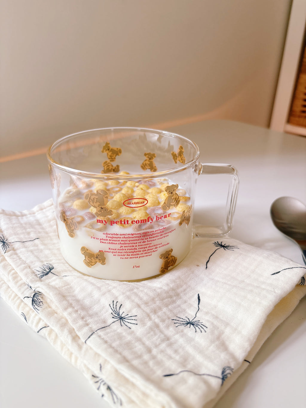 A clear glass cereal cup with a handle, filled with milk and cereal, resting on a white muslin cloth with a dandelion pattern. The cup features a red printed label that reads 'my petit comfy cup' and '1200 ml'.