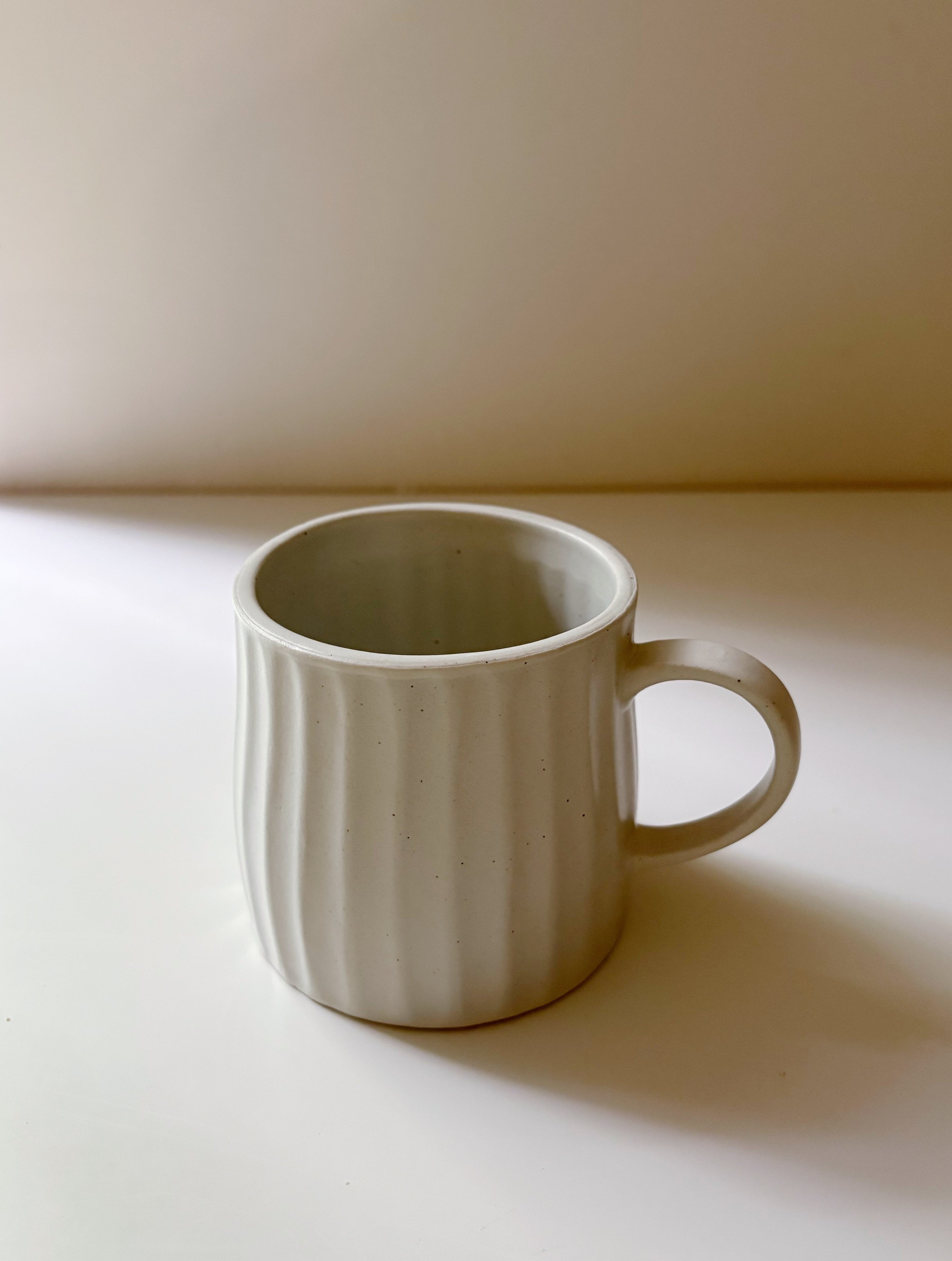 A light beige ceramic mug with a ribbed vertical texture and a C-shaped handle, sitting on a white surface with a warm beige background.