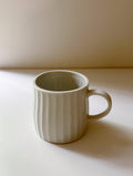 A light beige ceramic mug with a ribbed vertical texture and a C-shaped handle, sitting on a white surface with a warm beige background.