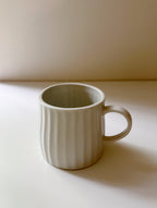 A light beige ceramic mug with a ribbed vertical texture and a C-shaped handle, sitting on a white surface with a warm beige background.