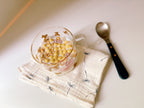 A clear glass mug filled with cereal and milk sits on a cream muslin cloth with black dandelion prints, next to a metal spoon with a black handle, on a warm off-white tabletop.