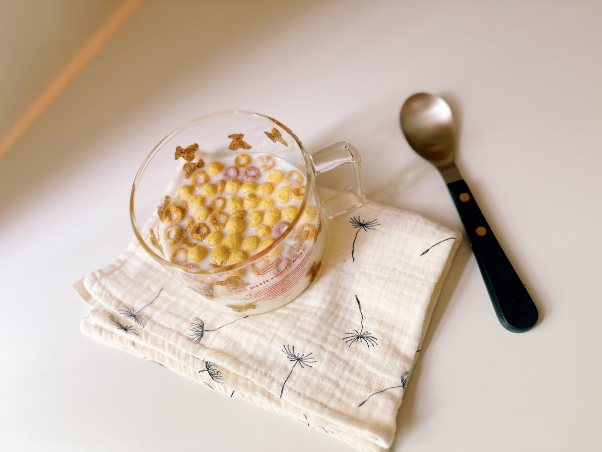 A clear glass mug filled with cereal and milk sits on a cream muslin cloth with black dandelion prints, next to a metal spoon with a black handle, on a warm off-white tabletop.