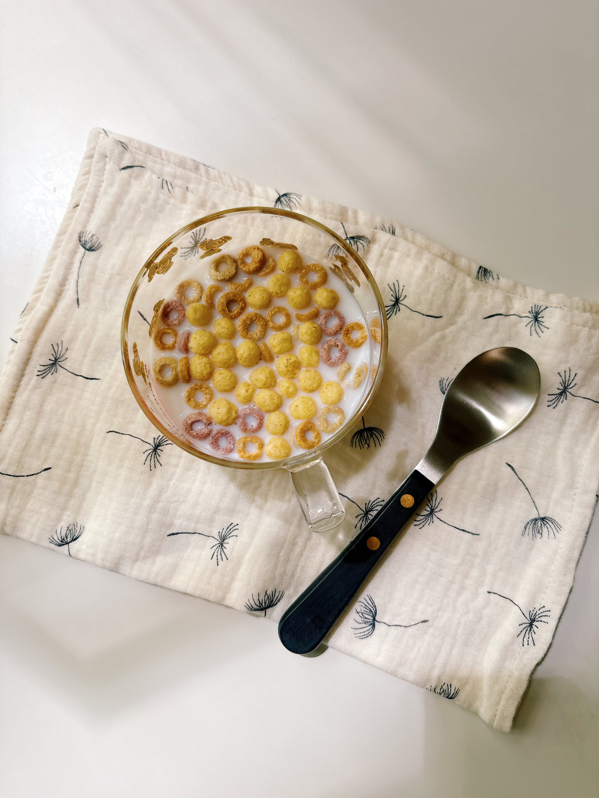 A clear glass mug filled with yellow and pink cereal rings in milk, placed on a cream cloth with a dandelion print, alongside a metal spoon with a dark handle on a white surface.