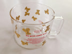 A clear glass cup with a handle, decorated with gold teddy bear illustrations and red text reading 'my petit comfy bear' and 'MARIE', photographed on a light neutral background.