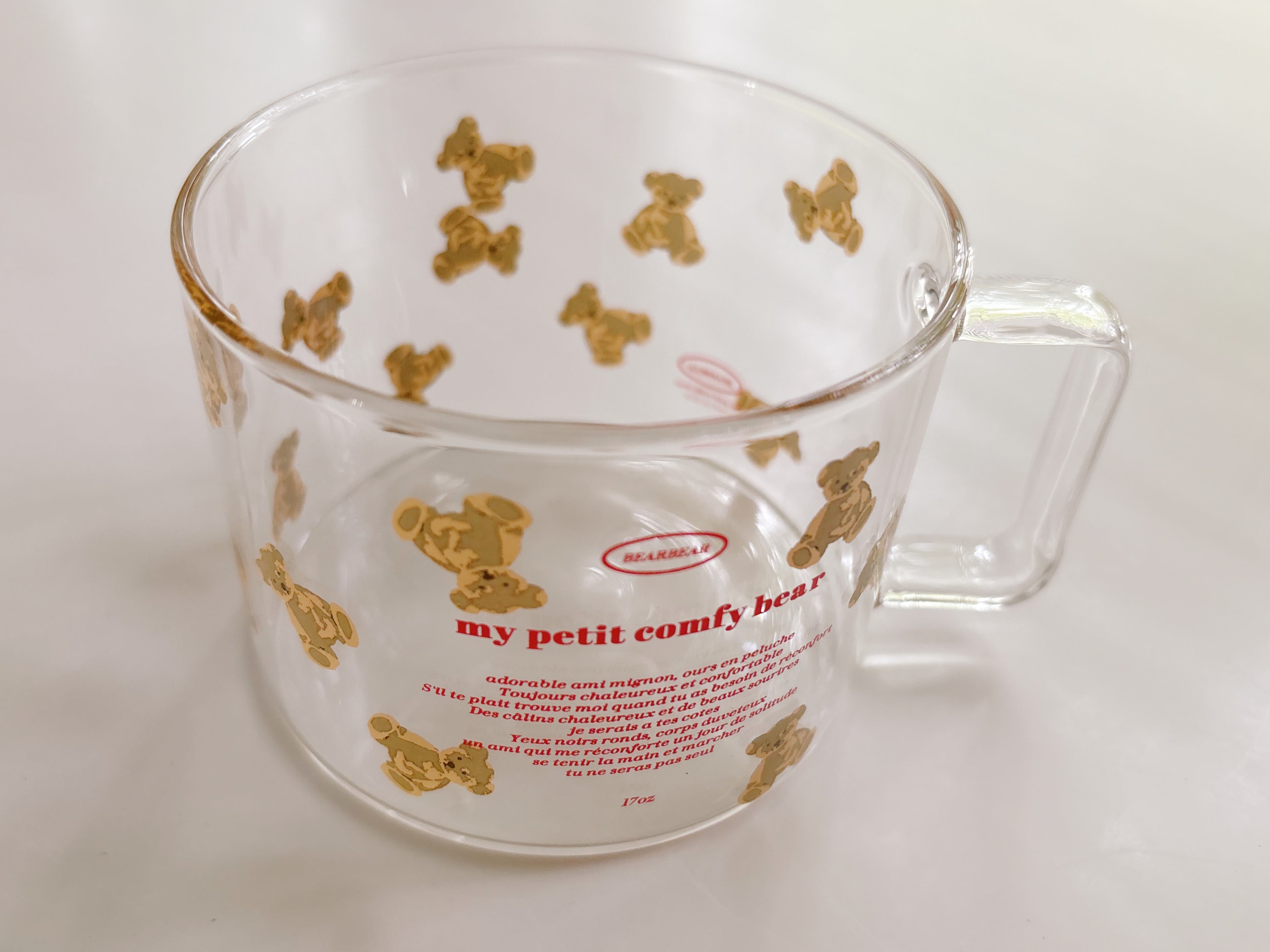 A clear glass cup with a handle, decorated with gold teddy bear illustrations and red text reading 'my petit comfy bear' and 'MARIE', photographed on a light neutral background.