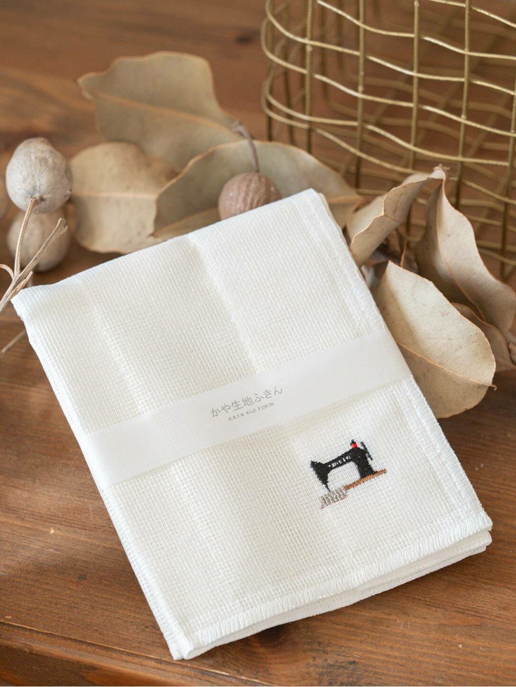 A folded white cotton handkerchief with a small black and red sewing machine logo and Japanese text, wrapped in a white paper band, displayed on a wooden surface with dried leaves and decorative props.