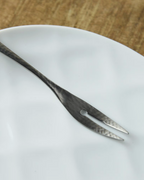 A silver metal serving fork with a hammered texture rests diagonally on a white ceramic plate, set against a warm wooden tabletop background.