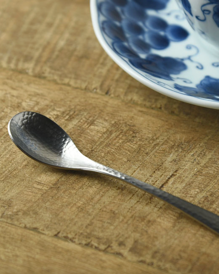 A small hammered metal spoon resting on a rustic wooden surface, with a blue and white patterned ceramic cup and saucer partially visible in the upper right.