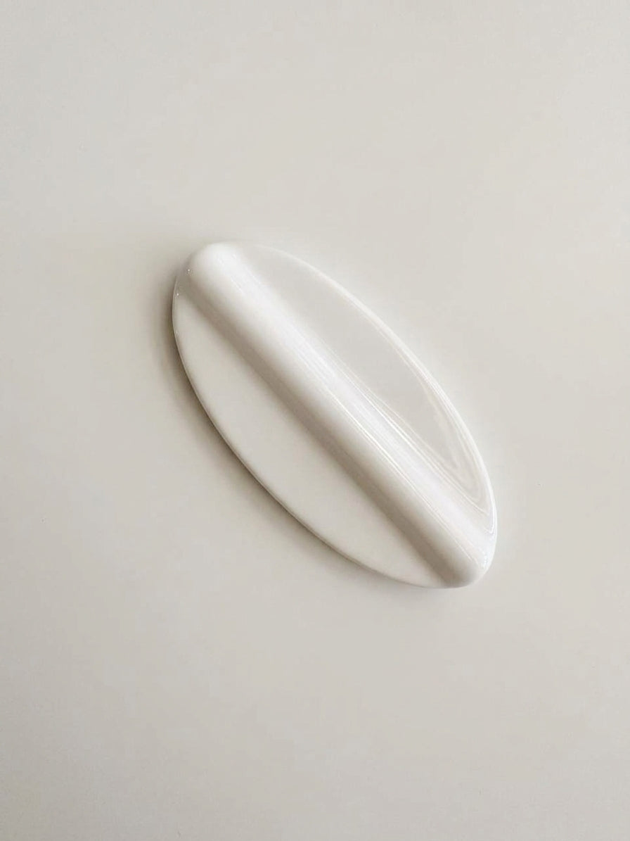 A glossy white, elongated oval-shaped spoon rest, possibly a decorative soap dish or a small sculptural vessel, shown on a plain light beige background.